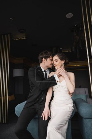 glücklicher Bräutigam im schwarzen Anzug mit Krawatte berührt Gesicht der charmanten Braut in weißem Brautkleid und Schmuck auf blauem Sofa in modernem Hotelzimmer gelehnt 