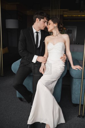 tierno novio en traje negro abrazando la cintura de la novia encantadora en vestido de novia blanco y joyas mientras se apoyan juntos en el sofá azul en la habitación de hotel moderna 