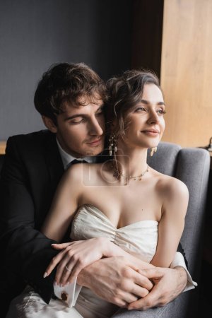 gut aussehender Bräutigam im schwarzen Anzug umarmt charmante Braut in weißem Kleid und luxuriösem Schmuck, während sie lächelt und zusammen auf bequemen Sesseln im Hotelzimmer sitzt 