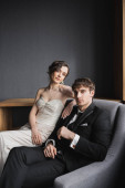 brunette woman in white wedding dress and luxurious jewelry sitting on comfortable armchair together with good looking groom in black suit in hotel room  Poster #654953356