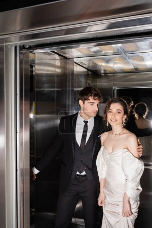 gut aussehender Bräutigam in formeller Kleidung, der Knopf drückt und fröhliche Braut im Brautkleid umarmt, während sie zusammen im Fahrstuhl des modernen Hotels steht 