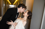 side view of happy groom in suit leaning towards charming bride in white wedding dress while smiling together in corridor of modern hotel, couple on honeymoon   Sweatshirt #654953480
