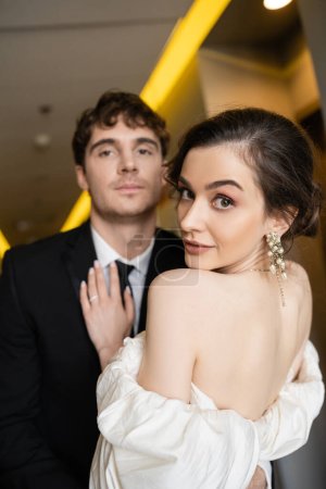 stunning and young bride in white wedding dress and earrings with pears standing near groom in black suit on blurred background in corridor of modern hotel, honeymoon  
