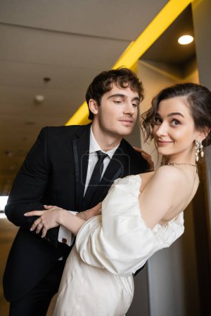 cheerful man in black suit leaning towards gorgeous bride in white wedding dress while smiling together and standing in corridor of modern hotel, honeymoon concept 