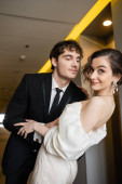 cheerful man in black suit leaning towards gorgeous bride in white wedding dress while smiling together and standing in corridor of modern hotel, honeymoon concept  t-shirt #654953542