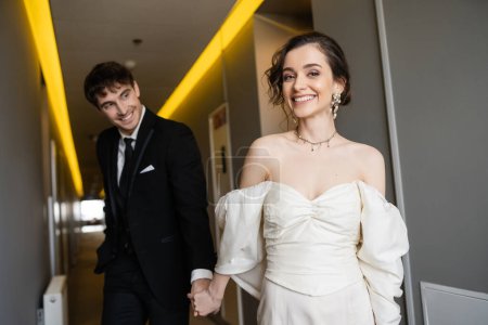 blurred and cheerful man in black suit holding hands with gorgeous bride in white wedding dress while smiling and walking together in corridor of modern hotel, honeymoon  