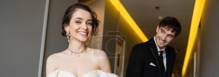blurred and cheerful man in black suit looking at gorgeous bride in white wedding dress while smiling and walking together in corridor of modern hotel, banner 