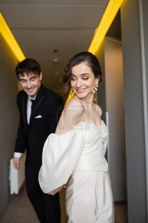 Photo for Delightful bride in white wedding dress holding hands with blurred and cheerful groom in black suit while smiling and walking together in hallway of modern hotel, happy newlyweds on honeymoon - Royalty Free Image