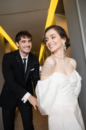 fröhlicher Bräutigam im schwarzen Anzug kitzelt entzückende Braut im weißen Hochzeitskleid, während sie lächelt und gemeinsam im Flur des modernen Hotels spaziert, glückliches Brautpaar auf Hochzeitsreise 