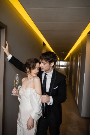 junger Bräutigam im schwarzen Anzug lehnt an der Wand und hält Flasche in der Nähe atemberaubende Braut mit einem Glas Champagner, während sie zusammen im Flur des modernen Hotels stehen, Brautpaar auf Hochzeitsreise 