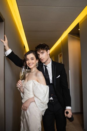 good looking groom in black formal wear leaning towards wall and holding bottle near happy bride with glass of champagne while standing together in hallway of modern hotel 