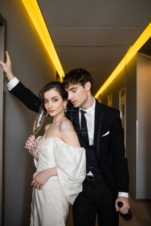 groom in black suit leaning towards wall and holding bottle near charming bride with glass of champagne while standing together in corridor of modern hotel  Mouse Pad 654953738