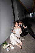 happy groom in black suit holding bottle and sitting near gorgeous bride with glass of champagne next to bridal bouquet and high heels on floor in corridor of modern hotel, newlyweds on honeymoon  mug #654953766
