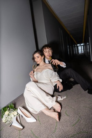 groom in black suit drinking from bottle and sitting near gorgeous bride with glass of champagne next to bridal bouquet and high heels on floor in hall of modern hotel 