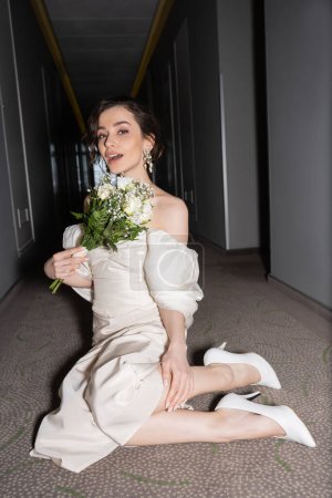 erstaunt junge Braut mit brünetten Haaren im weißen Hochzeitskleid blickt in die Kamera, während sie Brautstrauß mit Blumen in der Hand hält und auf dem Fußboden des Flurs im modernen Hotel sitzt 