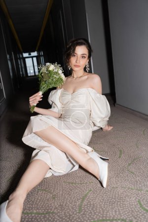 junge und brünette Braut im weißen Hochzeitskleid, die in die Kamera schaut, während sie den Brautstrauß mit Blumen in der Hand hält und auf dem Fußboden des Flurs im modernen Hotel sitzt 