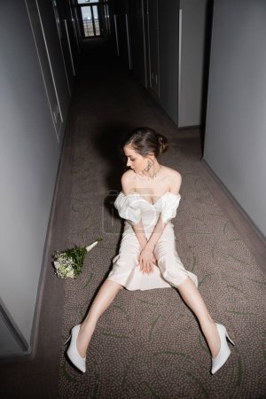 high angle view of young bride in white wedding dress and high heels looking at bridal bouquet with flowers while sitting on floor of hallway in modern hotel 