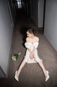 high angle view of young bride in white wedding dress and high heels looking at bridal bouquet with flowers while sitting on floor of hallway in modern hotel  puzzle #654953912