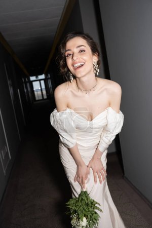 happy and brunette young bride in white wedding dress smiling while holding bridal bouquet with flowers and looking at camera in hall in modern hotel 