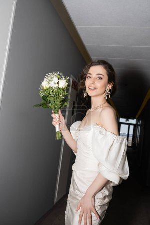 fröhliche und brünette junge Braut im weißen Kleid lächelnd, während sie den Brautstrauß mit Blumen in der Hand hält und in die Kamera im Saal des modernen Hotels schaut, schöne Braut am Hochzeitstag