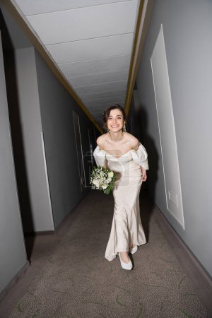 Photo for Full length of excited and brunette young bride in white wedding dress smiling while holding bridal bouquet with flowers and looking at camera in hall in modern hotel - Royalty Free Image