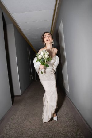 volle Länge der aufgeregten und brünetten Braut mit geöffnetem Mund, die im weißen Hochzeitskleid steht und einen Brautstrauß mit Blumen hält, während sie in die Kamera im Saal des modernen Hotels schaut 