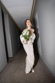 full length of excited and brunette bride with opened mouth standing in white wedding dress and holding bridal bouquet with flowers while looking at camera in hall in modern hotel  puzzle #654953962