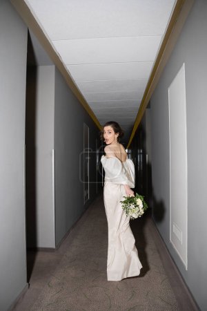 volle Länge der betäubten und brünetten Braut mit geöffnetem Mund, die im weißen Hochzeitskleid steht und einen Brautstrauß mit Blumen hält, während sie in die Kamera im Saal des modernen Hotels schaut 