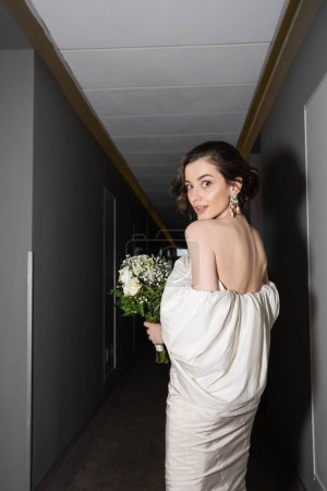 Photo for Stunned and brunette bride in white wedding dress holding bridal bouquet with flowers and looking at camera while standing in hallway of modern hotel - Royalty Free Image