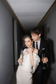 young and brunette bride in white wedding dress and handsome groom holding glasses of champagne while standing together in hallway of hotel, newlyweds on honeymoon  Stickers #654953998