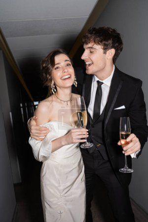 cheerful groom hugging young and brunette bride in white wedding dress and holding glasses of champagne while standing and smiling together in hallway of hotel, newlyweds on honeymoon 