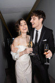 cheerful groom hugging young and brunette bride in white wedding dress and holding glasses of champagne while standing and smiling together in hallway of hotel, newlyweds on honeymoon  Tank Top #654954002