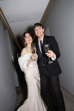 cheerful groom and young bride in white wedding dress and holding glasses of champagne while standing and smiling together in hallway of hotel, newlyweds on honeymoon 