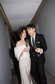 emotional groom hugging young and happy bride in white wedding dress and holding glasses of champagne while standing and smiling together in hallway of hotel  Poster #654954116