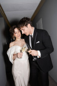 cheerful groom and brunette bride in white wedding dress and clinking glasses of champagne while standing and smiling together in hallway of hotel, newlyweds on honeymoon  mug #654954122