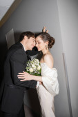 positive groom in black suit leaning towards wall near bride in white wedding dress holding bridal bouquet while standing together in hallway of modern hotel, newlyweds on honeymoon  puzzle #654954186
