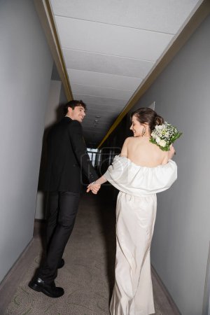 positive groom in black suit holding hands with bride in white wedding dress carrying bridal bouquet while walking together in hallway of modern hotel, newlyweds on honeymoon  tote bag #654954204