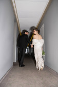 low angle view of groom in black suit holding hands with bride in white wedding dress carrying bridal bouquet while walking together in hallway of modern hotel, newlyweds on honeymoon  Longsleeve T-shirt #654954220