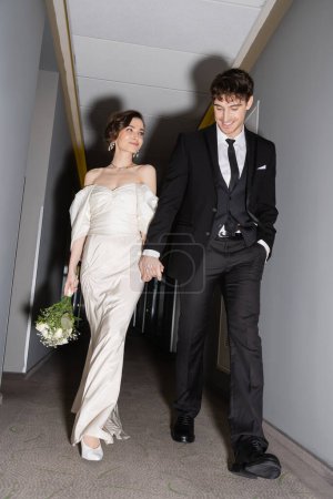 low angle view of cheerful groom in black suit posing with hand in pocket and holding hand of bride in white wedding dress carrying bridal bouquet while walking together in hall of modern hotel  Poster 654954236