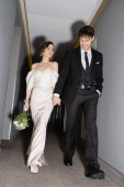 low angle view of cheerful groom in black suit posing with hand in pocket and holding hand of bride in white wedding dress carrying bridal bouquet while walking together in hall of modern hotel  t-shirt #654954236