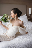 young bride in white dress and luxurious jewelry sitting on bed and smelling bridal bouquet with flowers while looking away in modern bedroom in hotel room on wedding day  puzzle #654954300