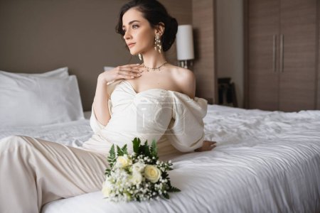 Bezaubernde Braut in weißem Kleid und luxuriösem Schmuck sitzt auf dem Bett neben Brautstrauß und schaut am Hochzeitstag im modernen Schlafzimmer im Hotelzimmer weg 