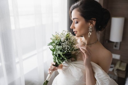 Foto de Vista lateral de la elegante novia joven en vestido blanco y joyas de lujo con pendientes de perlas que sostienen ramo de novia en la habitación de hotel moderna en el día de la boda - Imagen libre de derechos