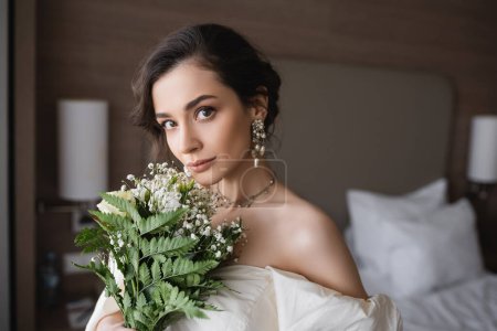 encantadora novia joven en vestido blanco y joyas de lujo celebración de ramo de novia con flores y mirando a la cámara en el dormitorio moderno en la habitación de hotel el día de la boda 
