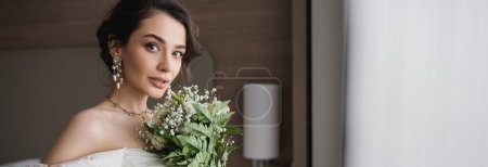 Charmante junge Frau in weißem Hochzeitskleid und luxuriösem Schmuck hält Brautstrauß mit Blumen und blickt in die Kamera im modernen Schlafzimmer im Hotelzimmer, Banner 