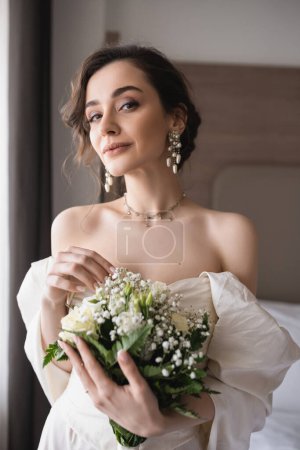 bezaubernde junge Frau in Brautkleid und luxuriösem Schmuck hält Brautstrauß mit Blumen in der Hand und blickt in die Kamera im modernen Schlafzimmer im Hotelzimmer am Hochzeitstag 