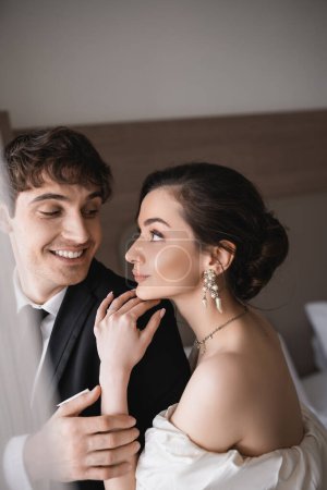 hübsche junge Braut in Schmuck und Brautkleid schaut fröhlichen Bräutigam in schwarzer Festkleidung an, während sie nach der Zeremonie im modernen Hotelzimmer zusammensteht 