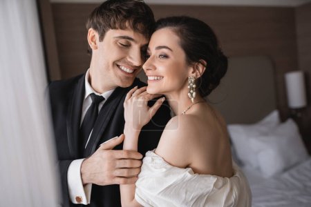 freudige junge Braut in Schmuck und Brautkleid umarmt Schulter eines fröhlichen Bräutigams in klassischer formeller Kleidung, während sie nach der Zeremonie im modernen Hotelzimmer zusammensteht 