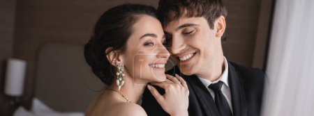 joyful young bride in jewelry hugging shoulder of cheerful groom in classic formal wear while standing together and smiling in modern hotel room after ceremony, banner 