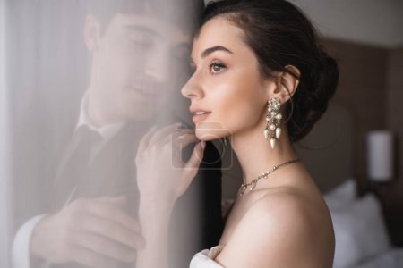 atemberaubende Braut in elegantem Schmuck und Brautkleid umarmt Schulter des Bräutigams in klassischer formeller Kleidung, während sie nach der Zeremonie im modernen Hotelzimmer hinter weißem Tüll zusammen steht 